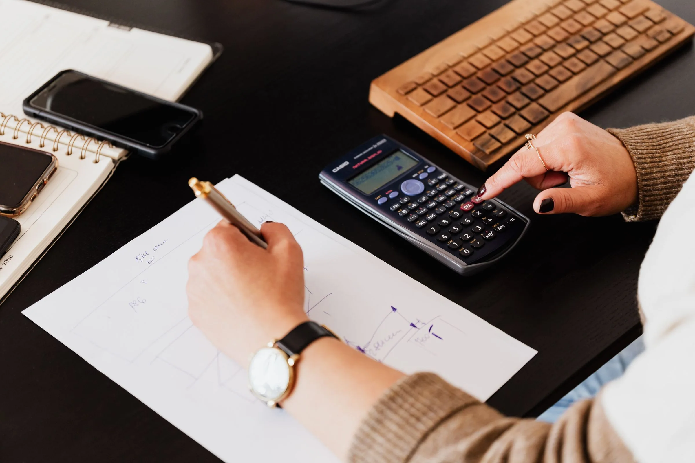 A person writing in a notebook while using a calculator at a desk